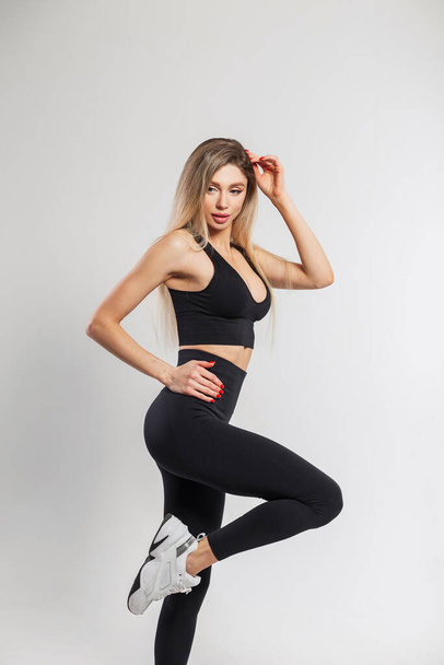 Красива атлетична леді з світлим волоссям в модному чорному спортивному одязі з верхнім одягом, легінси та кросівки позують на білому тлі в студії
 - Фото, зображення
