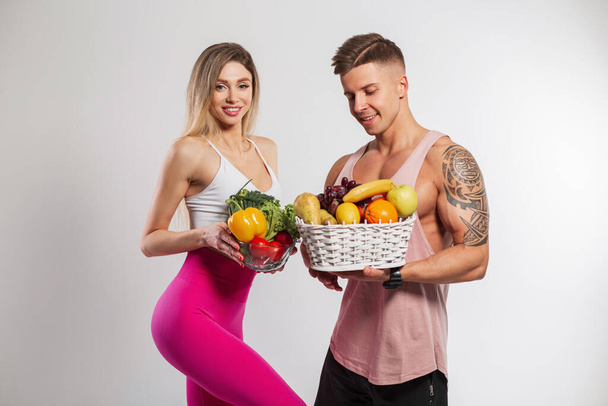 Тонкая семейная пара начала заниматься фитнесом, держа овощи и фрукты на белом фоне. Красивая стройная девушка и мускулистый мужчина с сырой диетической пищей - Фото, изображение