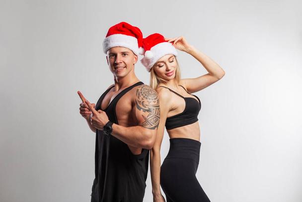 Beyaz arka planda kırmızı Noel Baba şapkalı şık spor giyimli şık bir çift. Yakışıklı, kaslı bir adam ve güzel bir fitness kadını. Yeni yıl ve spor yaşam tarzı - Fotoğraf, Görsel
