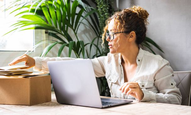 professional woman working at the desk with laptop and packages or letters to send. Concept of modern worker with online business. Young mature female people prepare shipment after sell. Home job - Photo, image