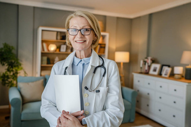 Ritratto di una donna caucasica bionda matura con occhiali medico stand medico durante la visita a casa sorriso felice guardando la fotocamera spazio copia sicuro - Foto, immagini