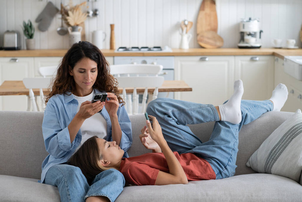 Addiction from internet woman mom and teen girl look at screen smartphone spend weekend time online. Thoughtful daughter lies on mother knees on home couch resting together immersive in cellphones - Zdjęcie, obraz