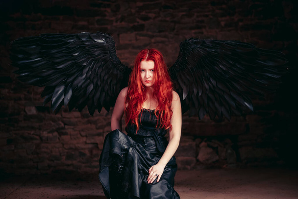 A girl with black angel wings sits on the floor in a dark room. Red-haired girl. - Фото, изображение