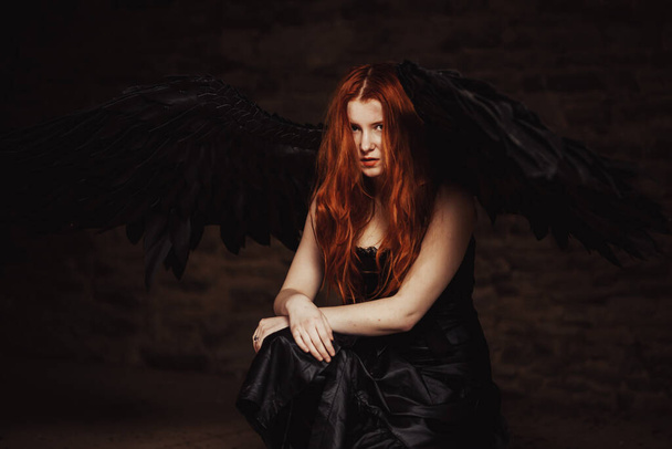 Dark angel with black wings on a stone dark background. Red-haired girl, witch sits on the floor. tinted - Fotoğraf, Görsel