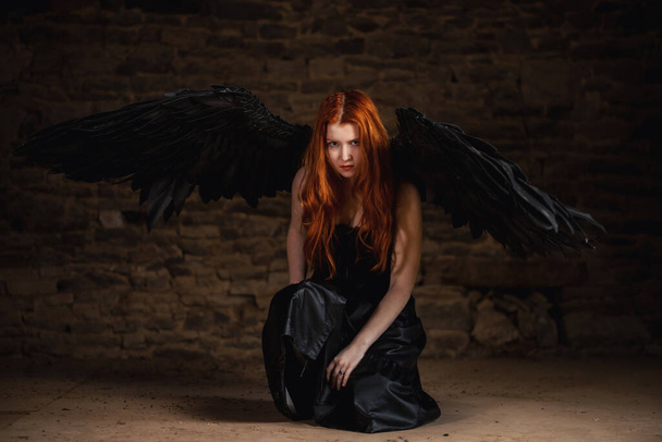 Dark angel with black wings on a stone dark background. Red-haired girl, witch sits on the floor. - Fotoğraf, Görsel