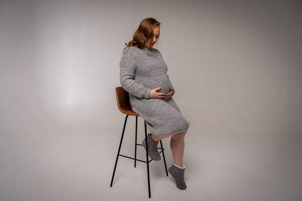 full length portrait of a pregnant woman resting sitting on a chair on a light background, excited in anticipation, full photo, space for text - Fotó, kép