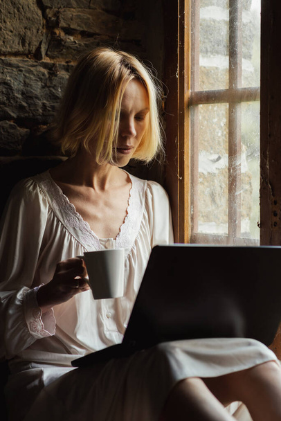 Woman with a laptop and a cup of coffee near the window. Middle age woman in a white shirt. Morning coffee. Freelancer. - Photo, Image