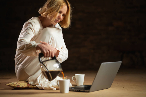 A woman pours coffee while sitting in front of a laptop. Empty room, dark background. Deadline, recycling, hard work freelancer concept. - Fotografie, Obrázek