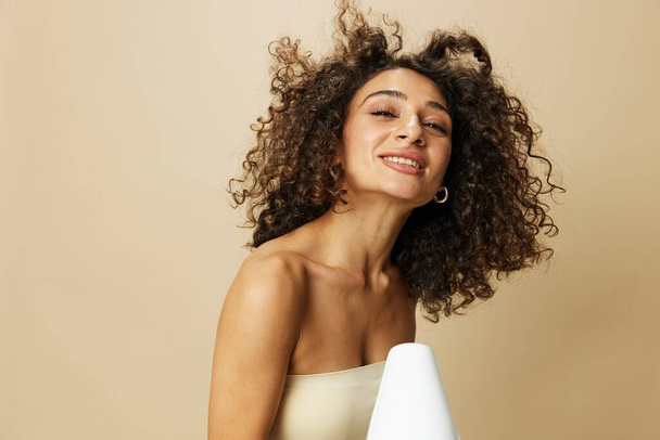 Donna asciuga i capelli ricci con asciugacapelli, casa batte prodotti per lo styling, sorriso su sfondo beige. Foto di alta qualità - Foto, immagini