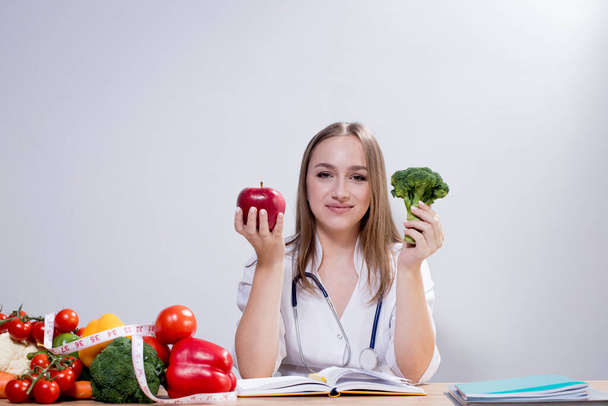 Positive young female nutritionist sitting at the table and holding broccoli and an apple in her hands. Concept of healthy eating. - Photo, Image