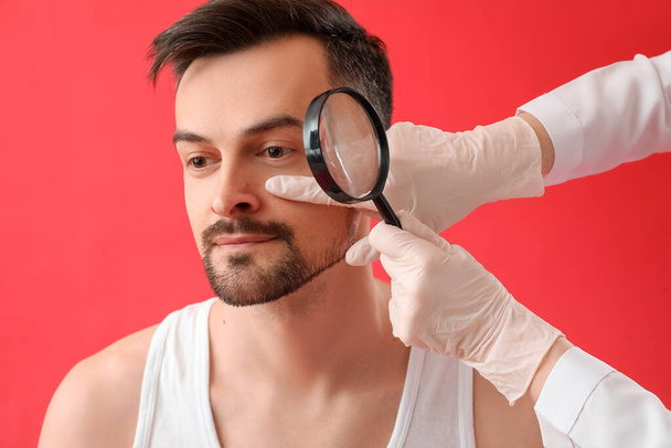 皮膚科医は、赤の背景に拡大鏡を持つ若い男のモルを調べます,クローズアップ - 写真・画像