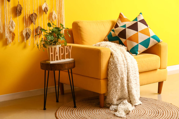 Acogedor sillón con cuadros y planta de interior en la mesa de centro cerca de la pared amarilla - Foto, Imagen