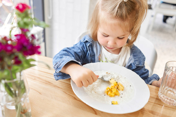 Een klein kind dat ontbijt in het café. Schattig meisje dat stilstaand water drinkt, rijstpap eet met mango. Geniet van het ontbijt. Hoge kwaliteit foto - Foto, afbeelding