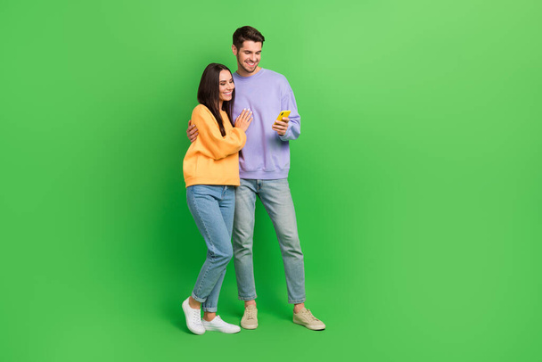 Full body foto van twee positieve partners knuffelen gebruik telefoon lege ruimte geïsoleerd op groene kleur achtergrond. - Foto, afbeelding
