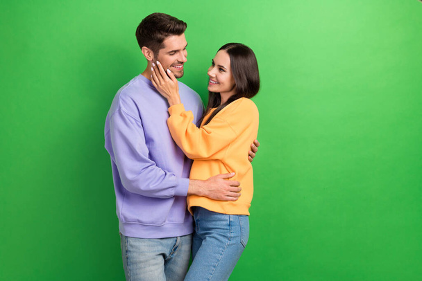 Portrait of two positive satisfied people cuddle hand touch face isolated on green color background. - 写真・画像