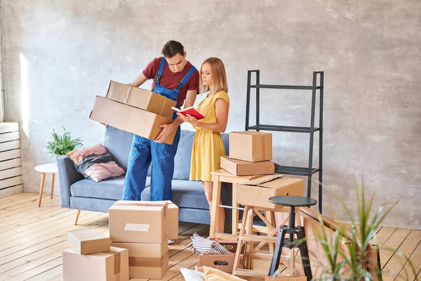 Delivery and moving day concept. The loader delivers the cargo, the house is full of boxes. Happy woman is new homeowner writing something on clipboard in room after moving day. High quality photo - Photo, Image