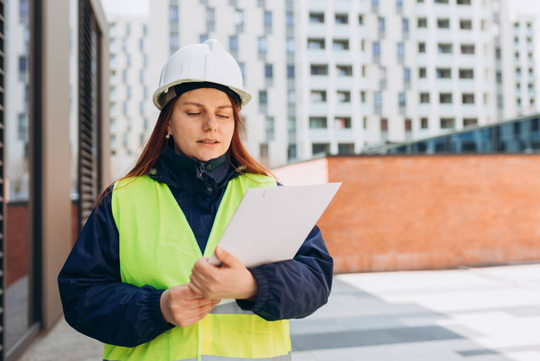 Architect with paper folder with documents at a construction site. Woman constructor wearing helmet and safety yellow vest. Women are planning new building project. - Photo, Image