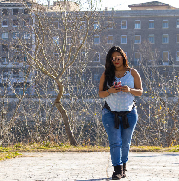 Beautiful latin woman walking in a park while writing a message on her cell phone, wearing blue jeans and blue t-shirt, with sunglasses. Trees and buildings in the background - Fotoğraf, Görsel