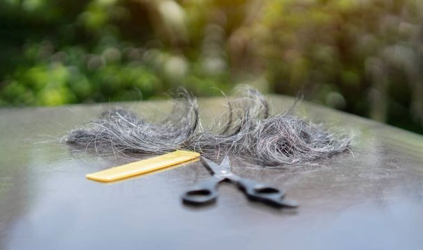 Tijeras de peluquería con peine amarillo y hebra de pelo rubio sobre mesa negra al aire libre con fondo natural. Enfoque selectivo. - Foto, Imagen