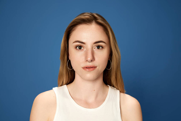 Portrait of young blonde girl posing, looking at camera with calm, attentive face against blue studio background. Student, education. Concept of youth, human emotions, facial expression - Photo, Image