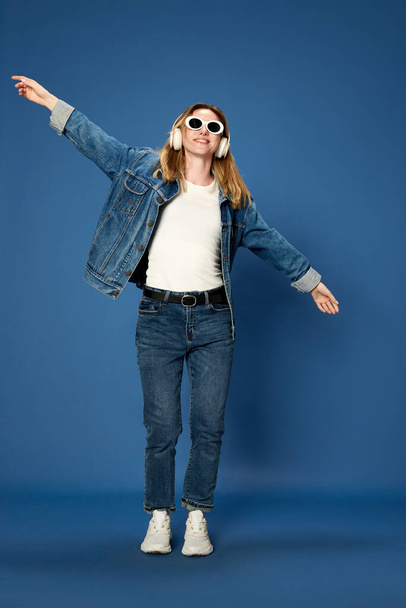 Full-length portrait of young girl in jeans outfit listening to music in headphones, smiling against blue studio background. Joy. Concept of youth, human emotions, facial expression - Zdjęcie, obraz