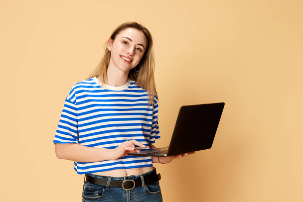 Portrait of young girl, student, employee in striped shirt styanding with laptop against yellow studio background. Success, win. Concept of youth, human emotions, facial expression, business and - Zdjęcie, obraz