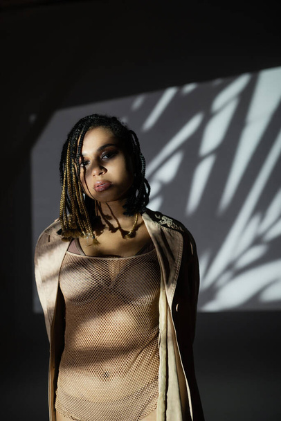 charming and sexy african american woman with stylish dreadlocks, in lingerie, net bodysuit and jacket looking at camera while posing in light on black and white background with white shadows - Zdjęcie, obraz