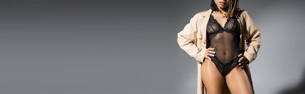 partial view of graceful and sexy african american woman in fashionable beige trench coat and black lace lingerie posing with hands on hips on grey background, banner - Foto, afbeelding