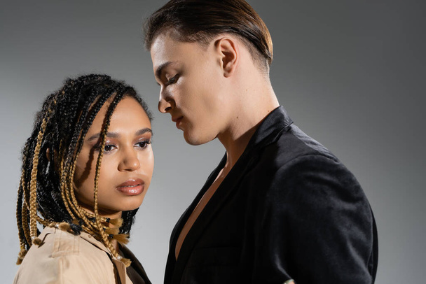 young and handsome man in black stylish blazer standing near charming african american woman with dreadlocks and smokey makeup looking away on grey background - Valokuva, kuva
