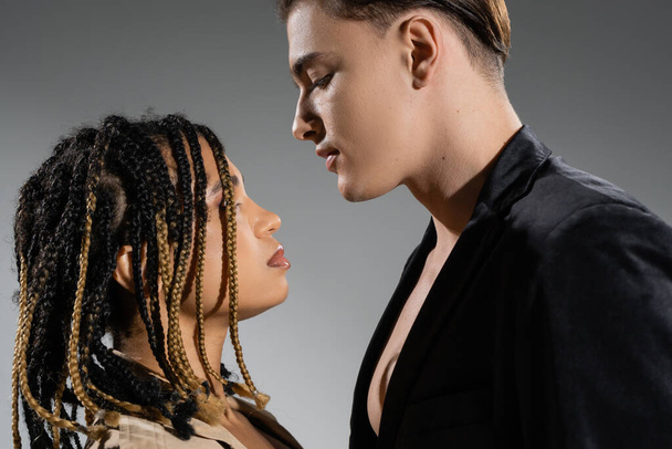 side view of sexy interracial couple, african american woman with stylish dreadlocks and young good looking man wearing black silk blazer on grey background - Foto, afbeelding