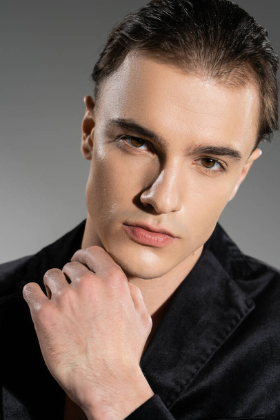 portrait of handsome and confident man with brunette hair posing in black and silk blazer and holding hand near chin while looking at camera on grey background - Фото, зображення