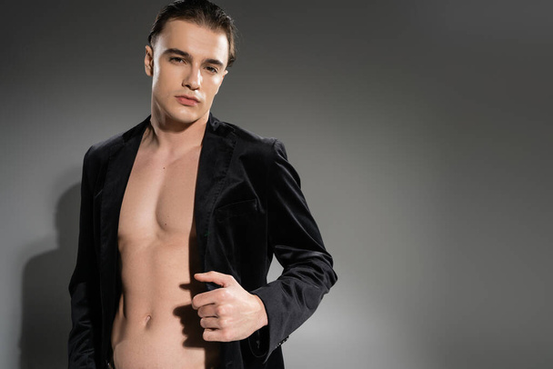 young, confident and good looking man with brunette hair, wearing black and silk blazer on shirtless muscular body while looking at camera on grey background - Photo, Image