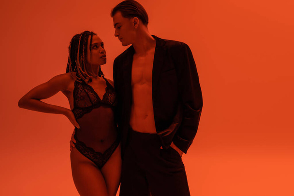 glamorous man in black blazer on shirtless body and sexy provocative african american woman with lace bodysuit looking at each other on orange background with red lighting effect - Fotografie, Obrázek