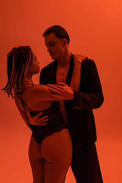 young and stylish man in black blazer hugging appealing african american woman with dreadlocks and sexy buttocks, wearing lace bodysuit on orange background with red lighting effect - Photo, Image