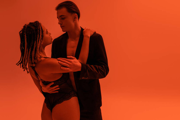 young and handsome man in stylish blazer embracing alluring african american man with sexy buttocks wearing lace bodysuit on orange background with red lighting effect - Zdjęcie, obraz