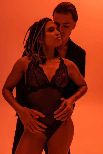 young self-assured man seducing sexy african american woman with dreadlocks and closed eyes, wearing black lace bodysuit on orange background with red lighting effect - Foto, imagen