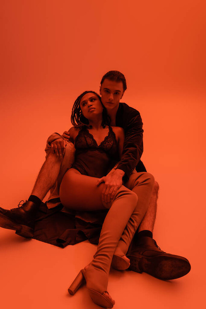 full length of young handsome man in blazer embracing sexy african american woman in black lace bodysuit and over knee boots while sitting on clothes on orange background with red lighting effect - Fotografie, Obrázek