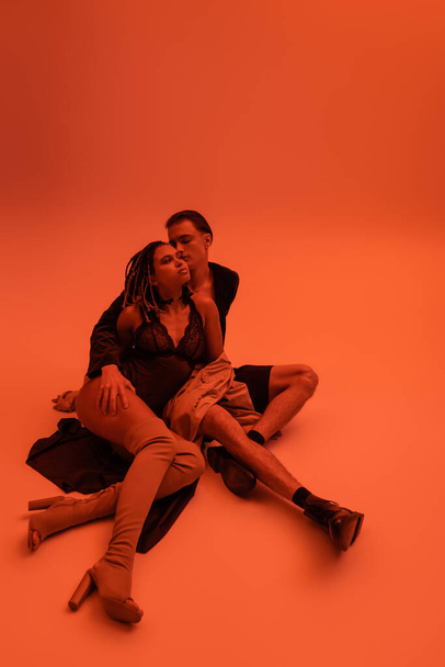 full length of passionate interracial couple, african american woman in black lace bodysuit with over knee boots, and young man sitting on clothes on orange background with red lighting effect - Photo, Image