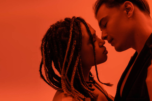 side view of young and sensual interracial couple, charismatic man and charming african american woman with dreadlocks kissing with closed eyes on orange background with red lighting effect - Fotografie, Obrázek