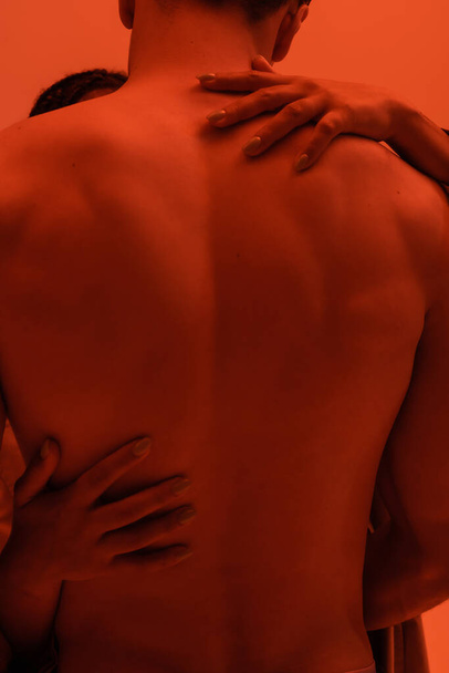 momento intimo di giovane e senza maglietta uomo vicino appassionato donna afroamericana abbracciando la sua schiena muscolare su sfondo arancione con effetto luce rossa - Foto, immagini