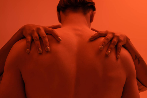 молодой мужчина без рубашки с мускулистой спиной рядом с страстной африканской женщиной обнимает свои плечи на оранжевом фоне с эффектом красного освещения - Фото, изображение