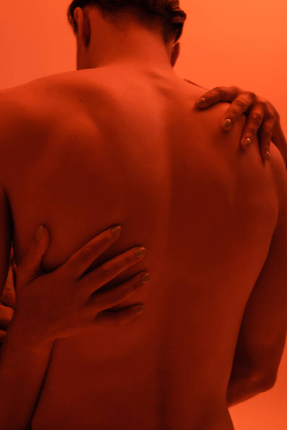 back view of young, shirtless and sexy man near impassioned african american woman embracing his muscular body on orange background with red lighting effect - Photo, Image