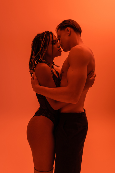 вид сбоку на межрасовую страстную пару, мужчину без рубашки в черных штанах с мускулистым туловищем и сексуальную африканскую женщину в кружевном костюме, обнимающую на оранжевом фоне с эффектом красного освещения - Фото, изображение
