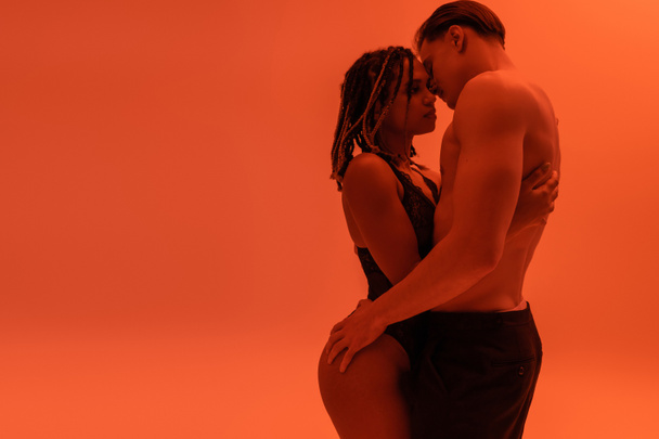side view of young shirtless man with muscular torso hugging sexy african american woman with dreadlocks, wearing black lace bodysuit on orange background with red lighting effect - Photo, Image