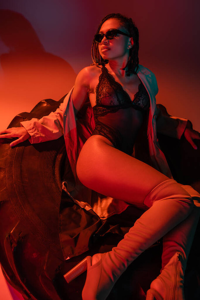 fashionable and sexy african american woman in sunglasses, black lace bodysuit, beige trench coat and over knee boots sitting on huge tire on grey background with orange lighting - Φωτογραφία, εικόνα