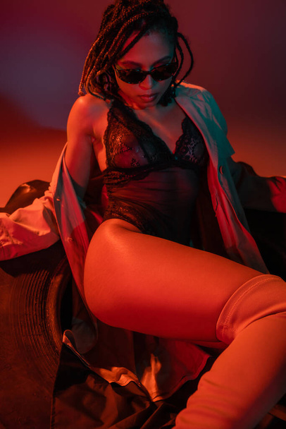 mujer americana africana atrevida y sexy en gafas de sol con estilo oscuro, traje de encaje negro y gabardina beige sentado sobre un enorme neumático sobre fondo gris con iluminación roja - Foto, imagen
