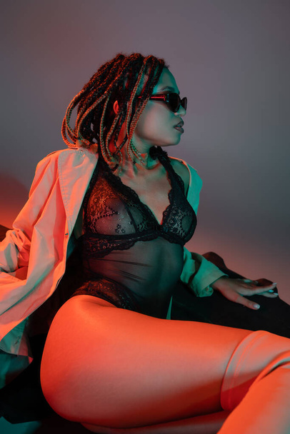 seductive african american woman with dreadlocks wearing dark sunglasses, black lace bodysuit and beige trench coat sitting on huge tire and looking away on grey background with red lighting - Foto, Bild