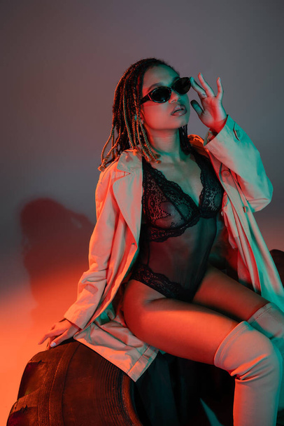glamorous and sassy african american woman adjusting dark stylish sunglasses while sitting on huge tire in black lace bodysuit and beige coat on grey background with red lighting - Фото, изображение