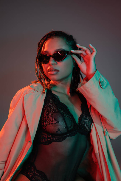 young and sexy african american woman with dreadlocks adjusting dark sunglasses while posing in black lace bodysuit and beige trench coat on grey background with red lighting - Photo, Image