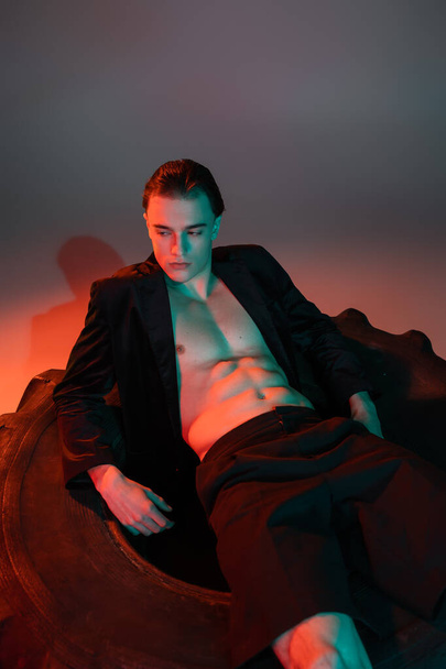 sexy and fashionable man with brunette hair, wearing black blazer on shirtless muscular body, sitting on huge tire and looking away on grey background with red lighting - Photo, Image
